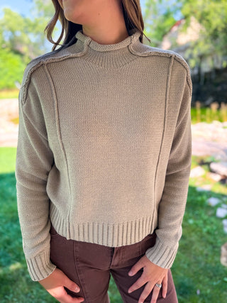 A woman wearing a beige ribbed knit mock neck sweater with shoulder detailing, paired with brown pants, standing outdoors on a grassy lawn.