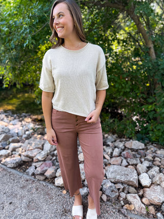 A woman standing outdoors in front of trees and rocks, wearing a short-sleeve cream knit top and dusty rose cropped pants with white slide sandals.