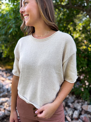 A woman wearing a short-sleeve knit top in a light cream or off-white color, standing outdoors in front of a blurred green foliage background.