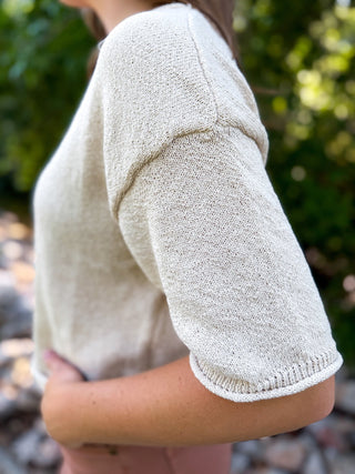 A close-up side view of a person wearing a short-sleeve knit top in a light oatmeal beige color, photographed outdoors with blurred green foliage in the background.