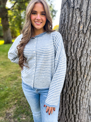 A woman wearing a blue and white striped knit cardigan and light blue jeans, standing outdoors next to a large tree trunk in a park setting.