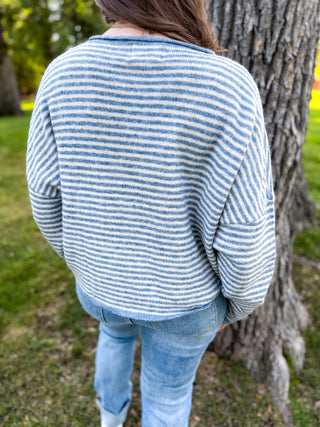 A person viewed from behind wearing a blue and white striped knit sweater and light blue jeans, standing outdoors next to a tree trunk in a grassy park setting.