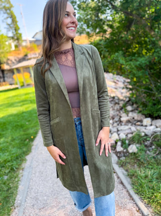 A woman wearing an olive green long-line suede jacket over a mauve lace top and blue jeans, standing on a gravel path outdoors with greenery and rocks in the background.