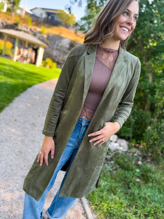 A woman wearing an olive green suede longline cardigan over a mauve lace-trim top and blue jeans, standing on a gravel path outdoors with greenery and a house in the background.