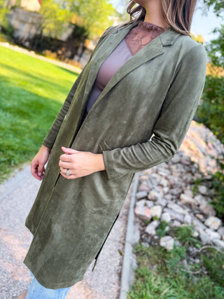 A woman wearing a long olive green suede-like open-front cardigan or duster over a mauve top, standing outdoors on a stone path with grass and trees in the background.