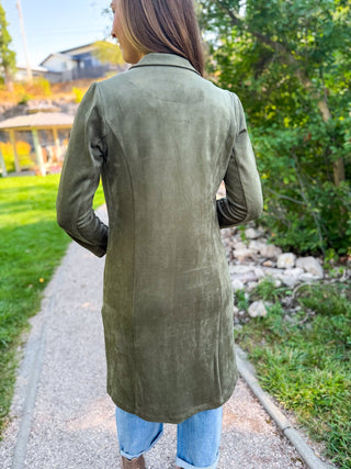 A woman viewed from behind wearing a long olive green suede-like blazer or tunic over light blue jeans, standing on a gravel path outdoors with greenery and a house in the background.