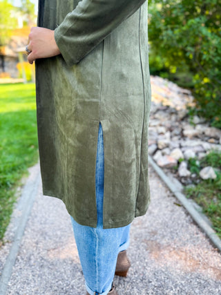 A person outdoors wearing a long olive green suede-like coat with a front slit, paired with light blue jeans and brown shoes, standing on a gravel path.