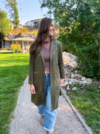 A woman walking outdoors on a gravel path wearing an olive green long-line suede jacket over a mauve lace top and light blue jeans.