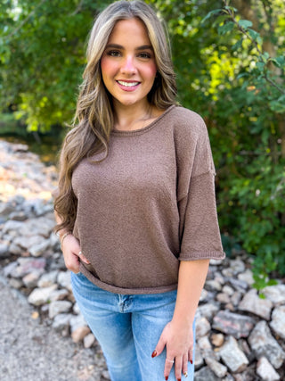 A woman with long wavy blonde hair stands outdoors wearing a brown knit crewneck sweater and blue jeans, with a background of green foliage and rocks.
