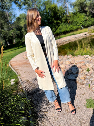 A woman stands outdoors on a gravel path near a pond, wearing a long cream-colored open-front cardigan over a black top and light blue jeans.