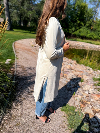 A woman wearing a long-sleeve cream linen-blend tunic top over blue jeans, standing on a gravel path in a sunny outdoor garden setting.