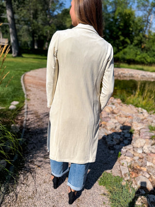 A woman viewed from behind wearing a long cream-colored suede or nubuck blazer over blue jeans, standing on a gravel path in a sunny outdoor garden setting.