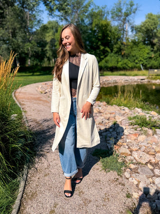 A woman stands on a gravel path outdoors near a pond, wearing a light cream open-front coat over a black lace-trim top, blue jeans, and black heeled sandals.