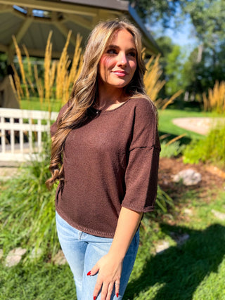 A woman wearing a brown knit top and light blue jeans standing outdoors in a garden setting with a house and trees in the background.