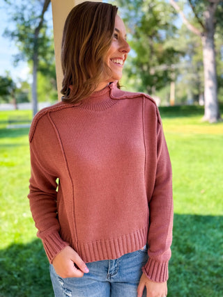 A woman wearing a dusty rose ribbed knit sweater with shoulder epaulets, paired with blue jeans, standing outdoors in a sunny park setting.