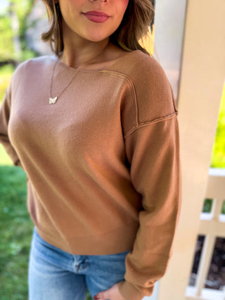A woman wearing a tan crewneck knit sweater and blue jeans, standing outdoors near a porch post.