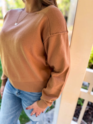 A woman wearing a ribbed crewneck sweatshirt in a warm camel brown color, paired with light blue jeans, standing outdoors next to a wooden post.