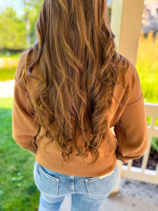 A woman viewed from behind wearing a camel brown long-sleeve knit top and light blue jeans, showcasing the back of the sweater with its relaxed fit and ribbed hem.