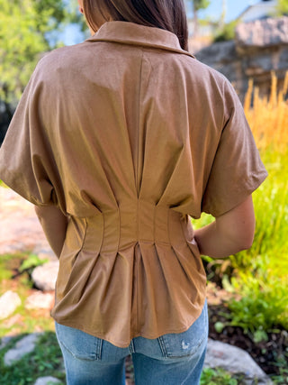 A person viewed from behind wearing a tan short-sleeve collared top with gathered detailing at the back and blue jeans, standing outdoors in a garden setting.