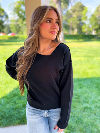 A woman with long light brown hair wearing a black long-sleeve top and light blue jeans, standing outdoors in a grassy park area.