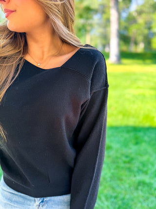 A woman wearing a black long-sleeve knit top with a wide scoop neckline, paired with light blue jeans, photographed outdoors against a blurred green park background.