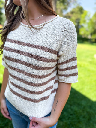A woman wearing a cream knit short-sleeve top with horizontal brown stripes, photographed outdoors on a grassy lawn.
