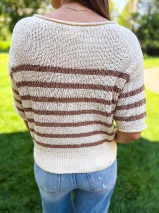 A person standing outdoors on grass, shown from the back wearing a cream knit sweater with horizontal brown stripes and light blue jeans.