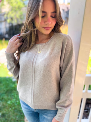 A woman wearing a light beige knit sweater and blue jeans, standing outdoors on a porch with green grass and trees in the background.