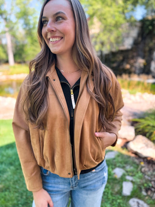 A woman wearing a tan suede zip-up jacket over a black top and blue jeans, standing outdoors in a garden setting.