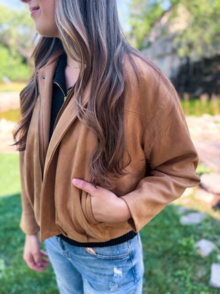 A woman wearing a tan faux-leather jacket, blue jeans, and a black top, standing outdoors in a grassy area with trees in the background.