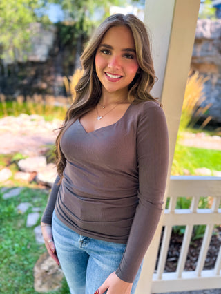A woman with long brown hair stands outdoors wearing a fitted, long-sleeve ribbed brown top and light blue jeans, posing in front of a porch post and garden background.
