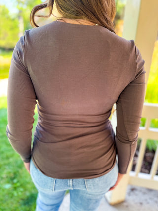 A woman viewed from behind wearing a fitted, long-sleeve brown ribbed knit top and light blue jeans, standing outdoors near a porch post.