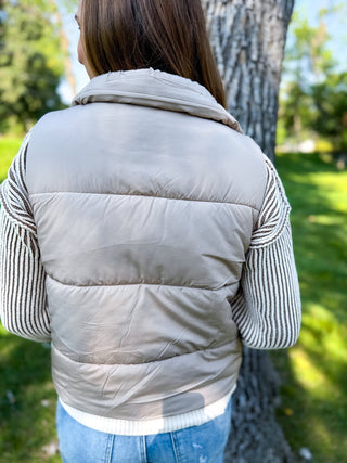 A woman viewed from behind wearing a light taupe-beige puffer vest over a striped long-sleeve top and blue jeans, standing outdoors next to a tree.