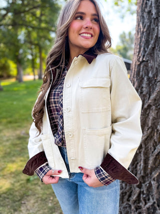 A woman wearing a cream-colored utility jacket with dark brown corduroy collar, cuffs, and plaid button-down shirt underneath, standing outdoors next to a tree.