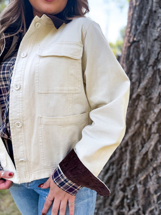 A person wearing a cream-colored canvas utility jacket with dark brown corduroy collar and cuff trim, layered over a plaid shirt and blue jeans, standing outdoors next to a tree trunk.