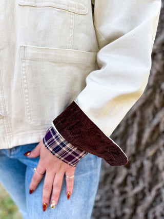 Close-up of a person wearing a cream canvas jacket with a dark brown corduroy cuff and a purple plaid lining, paired with blue jeans.