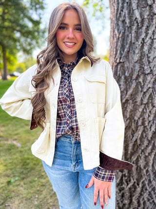 A woman wearing a cream-colored button-up utility jacket with brown corduroy cuffs, layered over a plaid shirt and blue jeans, standing outdoors next to a large tree trunk.