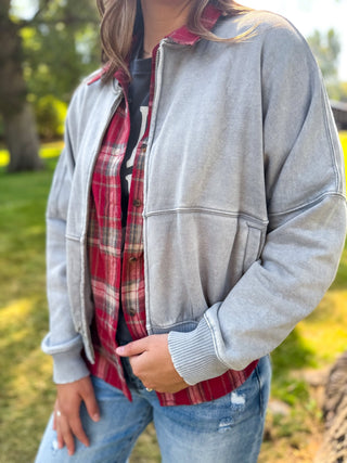 A person outdoors wearing a light gray oversized zip-front jacket over a red and black plaid flannel shirt, paired with blue jeans.