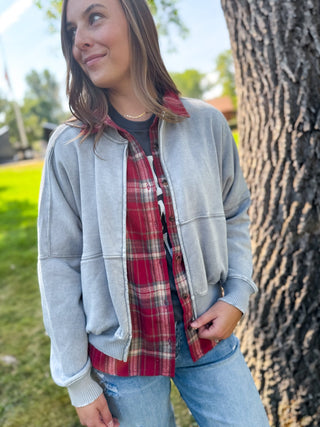 A woman wearing a light gray oversized zip-up jacket over a red and black plaid flannel shirt, paired with blue jeans, standing outdoors next to a large tree trunk.