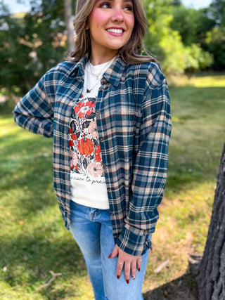 A woman standing outdoors in a grassy park area wearing a blue and beige plaid button-up shirt over a white graphic t-shirt and blue jeans.
