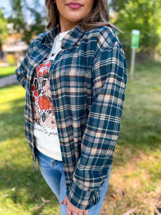 A woman wearing a teal and beige plaid flannel shirt over a white graphic t-shirt and blue jeans, standing outdoors on a grassy lawn.
