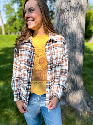A woman standing outdoors in front of a tree, wearing a yellow graphic t-shirt under an open long-sleeve plaid button-up shirt, paired with light blue jeans.