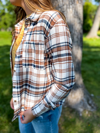 A woman wearing a long-sleeve plaid button-up shirt in brown, white, and navy, paired with blue jeans, standing outdoors in front of a tree.