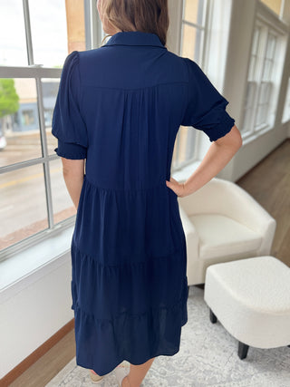 Woman wearing a navy blue dress in a room with a white chair and window.