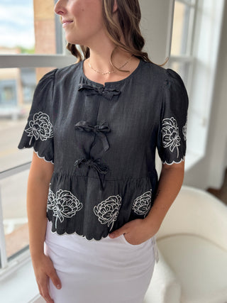 Woman wearing a black blouse with white floral embroidery in an indoor setting.
