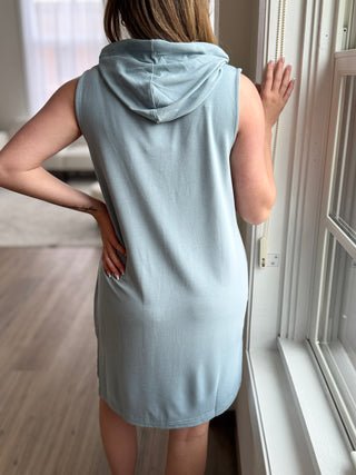 Person wearing a light blue sleeveless hoodie standing by a window.