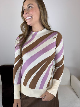 A woman wearing a long-sleeve knit sweater with diagonal wavy stripes in brown, lavender purple, and cream, standing in a brightly lit indoor setting.