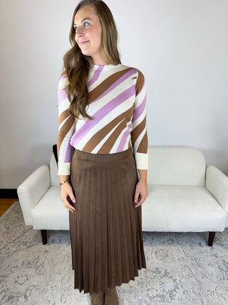 A woman modeling a striped knit sweater with brown, purple, and cream diagonal stripes, paired with a brown pleated midi skirt, standing in front of a white sofa.