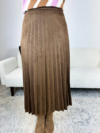A brown pleated faux-suede midi skirt worn by a model, paired with a striped top and brown ankle boots, set against a white armchair and patterned rug background.