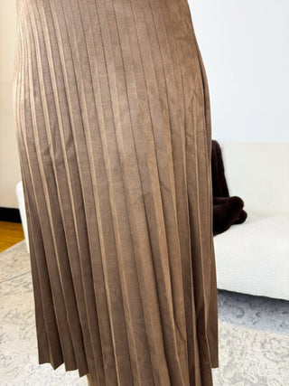 A close-up of a taupe brown pleated skirt, showing the texture and folds of the fabric, with a white sofa in the background.